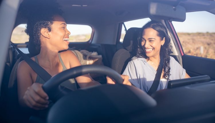 Young beautiful friends laughing while traveling in car during a road trip. Two multiethnic women driving in car and having fun. Two attractive girls talking together while driving in a car through the countryside on a sunset.