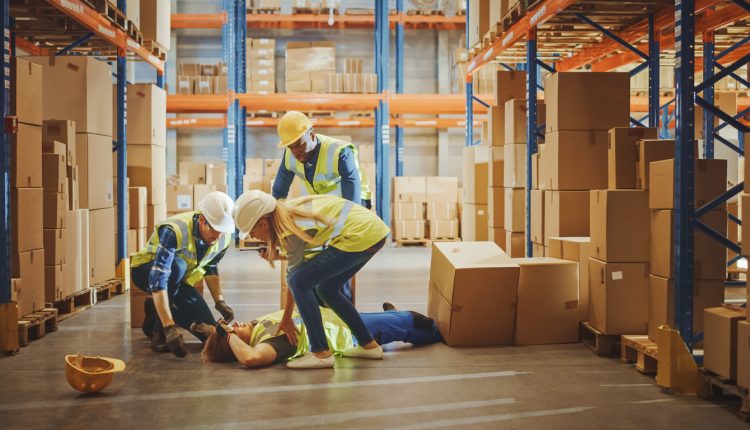 Warehouse Worker Has Work Related Accident Falls while Trying to Pick Up Cardboard Box from the Shelf. Colleagues Call for Help and Medical Assistance. Injury at Work.