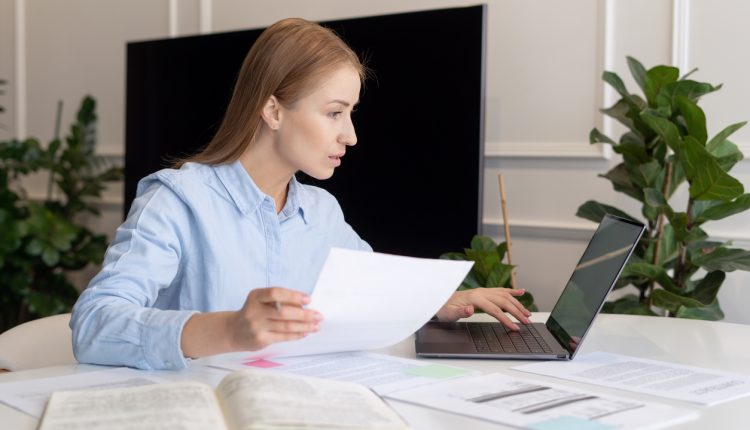 Side view of busy woman with documents in hands dealing with utility bills payment online. Usage of laptop to verify financial report results. Home office concept