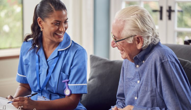Senior Man At Home Talking To Female Nurse Or Care Worker In Uniform Making Notes In Folder