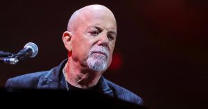 Billy Joel performing live on stage at the piano, looking emotional under stage lighting, with a microphone visible