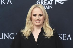 Rebel Wilson poses for photos at a formal event, wearing a black outfit with her long blonde hair styled in loose waves, standing in front of a branded backdrop that partially reads “GREAT BRITAIN” and “SEA.”