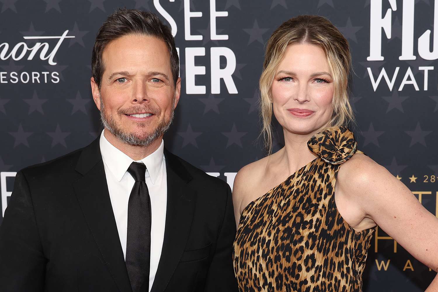 A close-up, half-body shot of actor Scott Wolf standing next to his estranged wife, Kelley Wolf, as they pose on a red carpet. Scott is on the left, wearing a black suit with a white shirt and black tie, and has a beard. Kelley is on the right, smiling and wearing a one-shoulder, leopard-print dress with a floral embellishment on the shoulder.