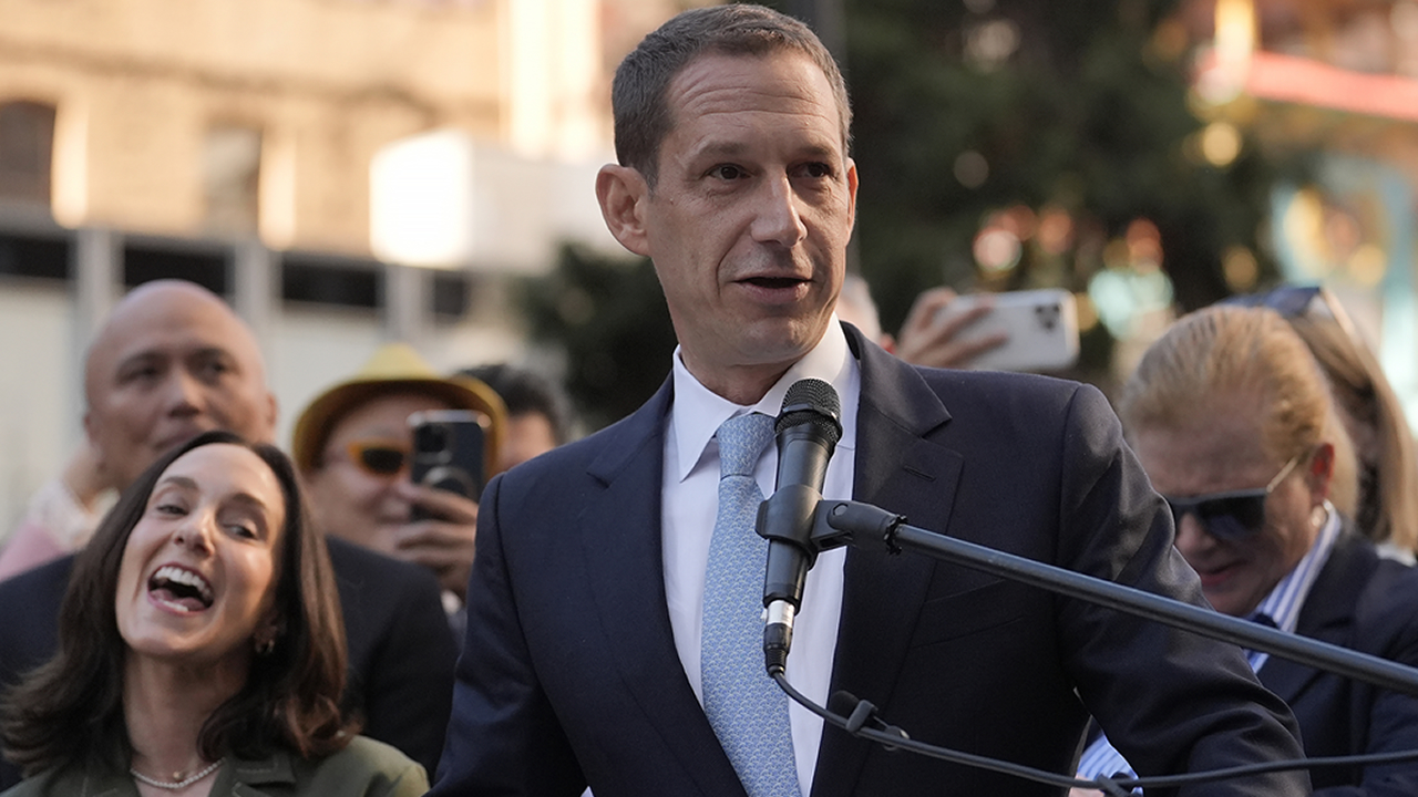 San Francisco Mayor Daniel Lurie speaks at a public event, addressing a crowd outdoors with a microphone, as people behind him listen and take photos.