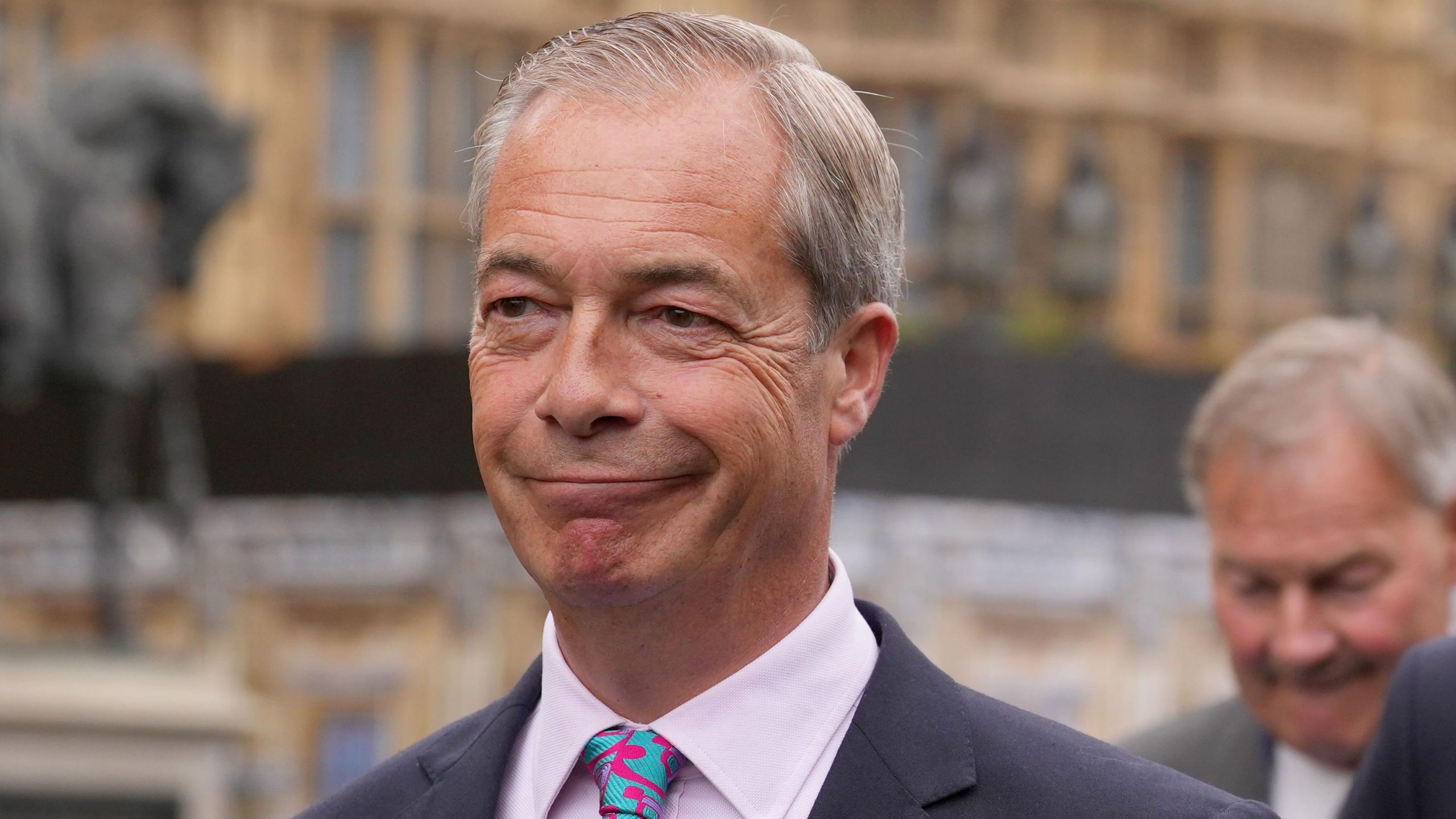 A middle-aged man in a blue suit and patterned tie looks directly at the camera with a serious expression, standing outdoors among other people