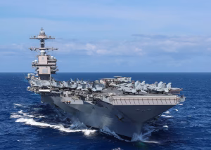 The USS Gerald R. Ford, the world’s largest aircraft carrier, sails through the Caribbean Sea with fighter jets and support aircraft visible on deck under clear blue skies.