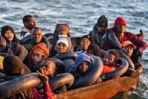 A group of migrants in a crowded wooden boat crossing the sea toward Europe, carrying inner tubes for safety during the Channel crossing.