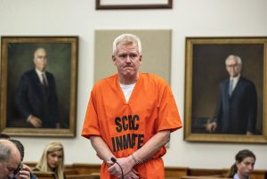 Alex Murdaugh stands in a courtroom wearing an orange South Carolina Department of Corrections inmate jumpsuit with his hands cuffed, flanked by portraits of former legal figures on the wall behind him.