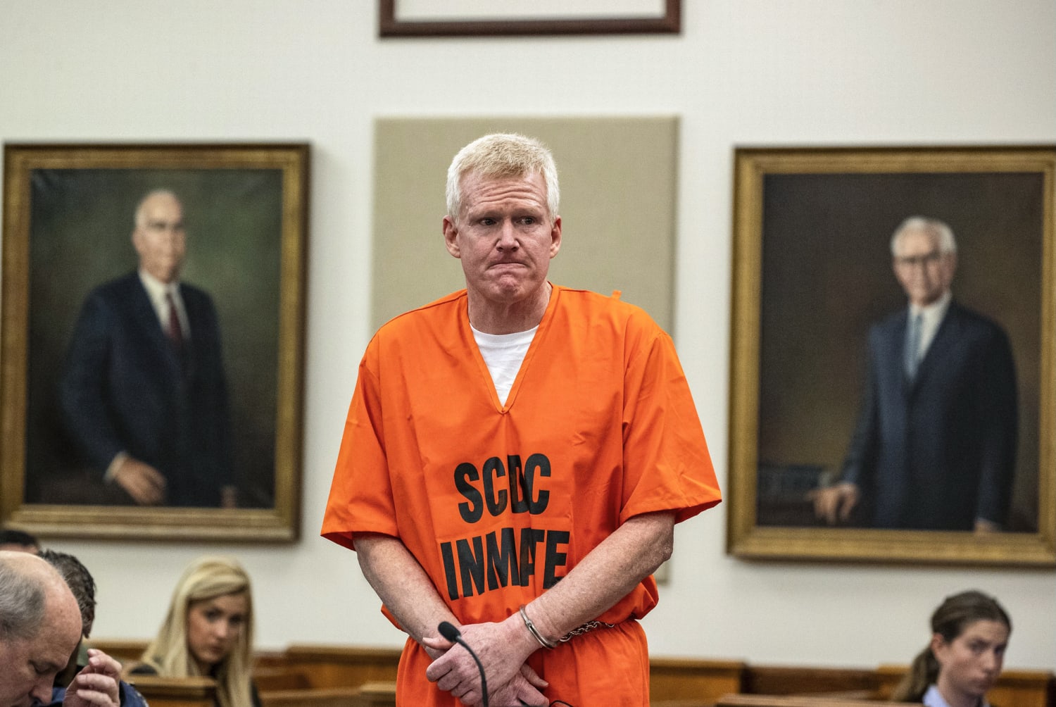 Alex Murdaugh stands in a courtroom wearing an orange South Carolina Department of Corrections inmate jumpsuit with his hands cuffed, flanked by portraits of former legal figures on the wall behind him.