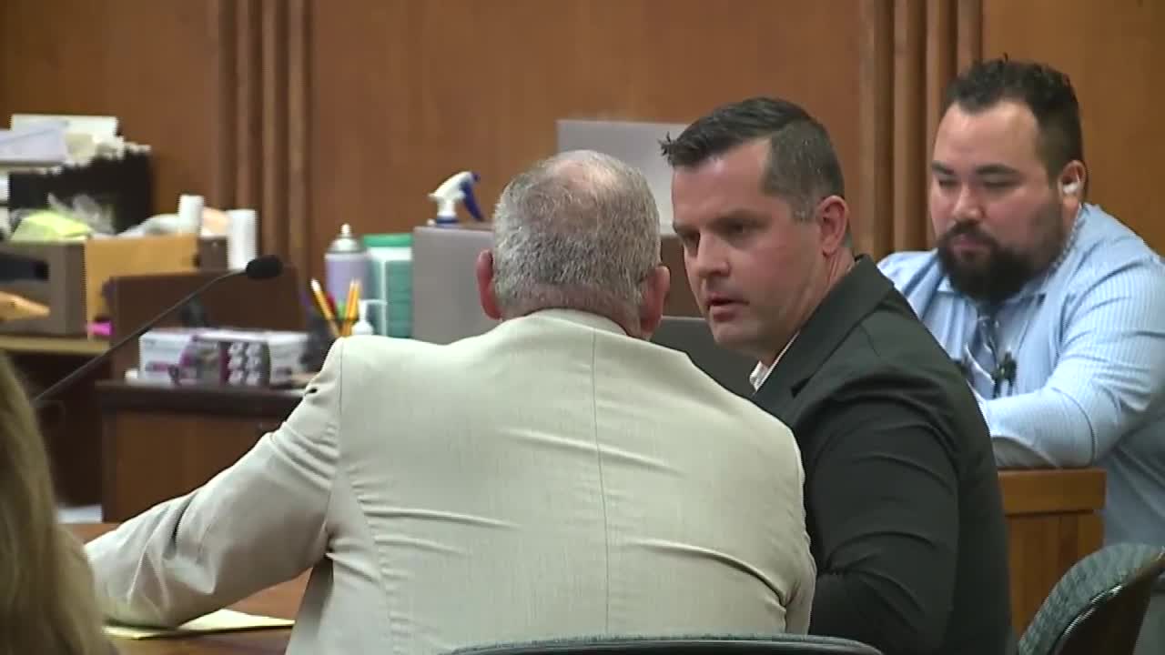 A man in a black jacket speaking with his defense attorney in a courtroom setting, with another man observing nearby.