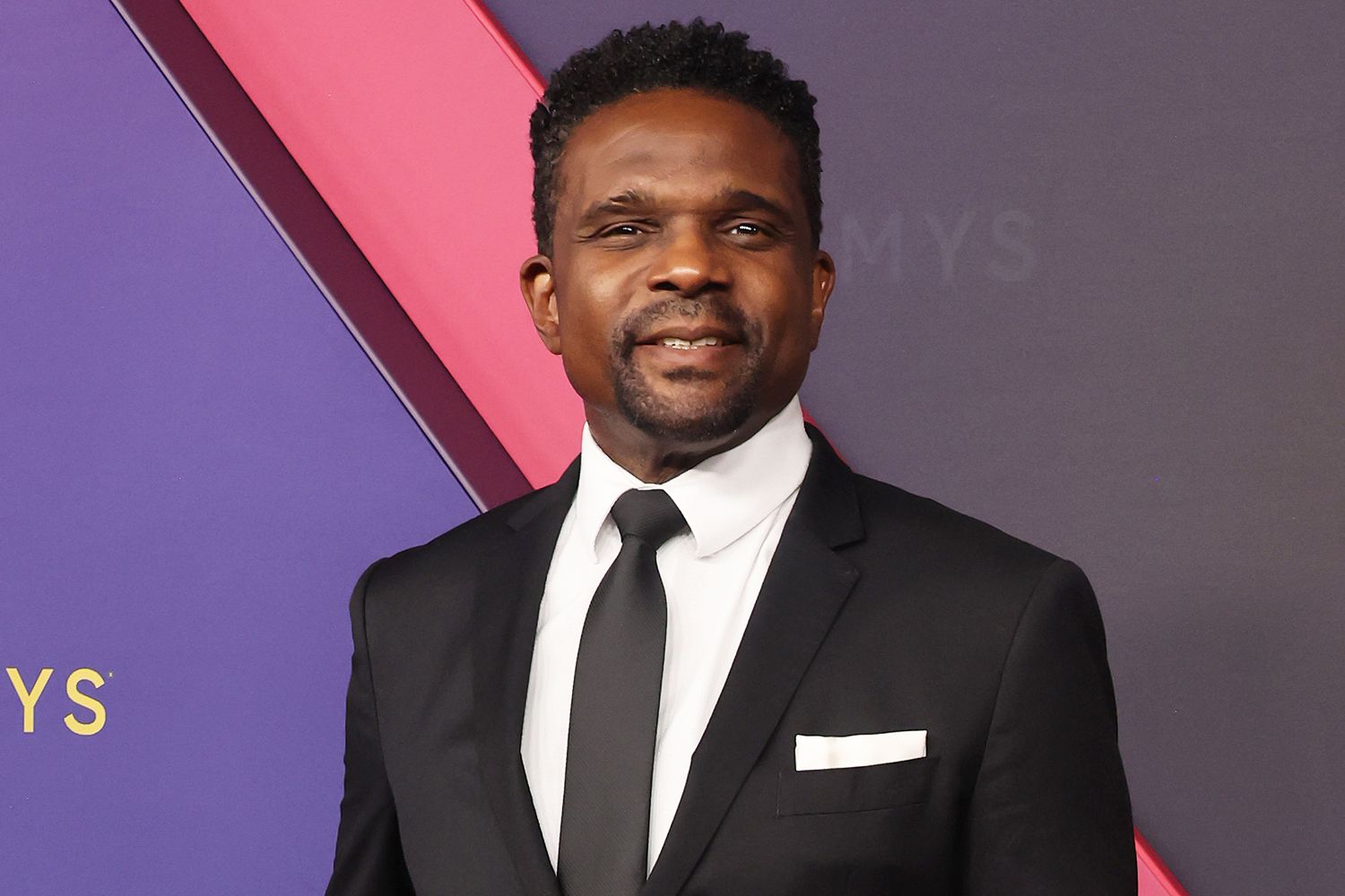 Darius McCrary wearing a black suit and tie at a red carpet event in Los Angeles, smiling for photographers.