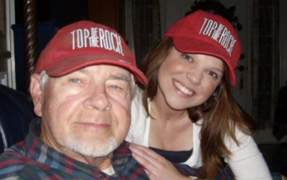 Amy Duggar King smiling next to her grandfather, Jimmy Lee Duggar, both wearing matching red "TOP THE ROCK" baseball caps.
