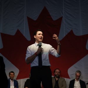 Justin Trudeau speaks on stage in front of a large Canadian flag during a public event.