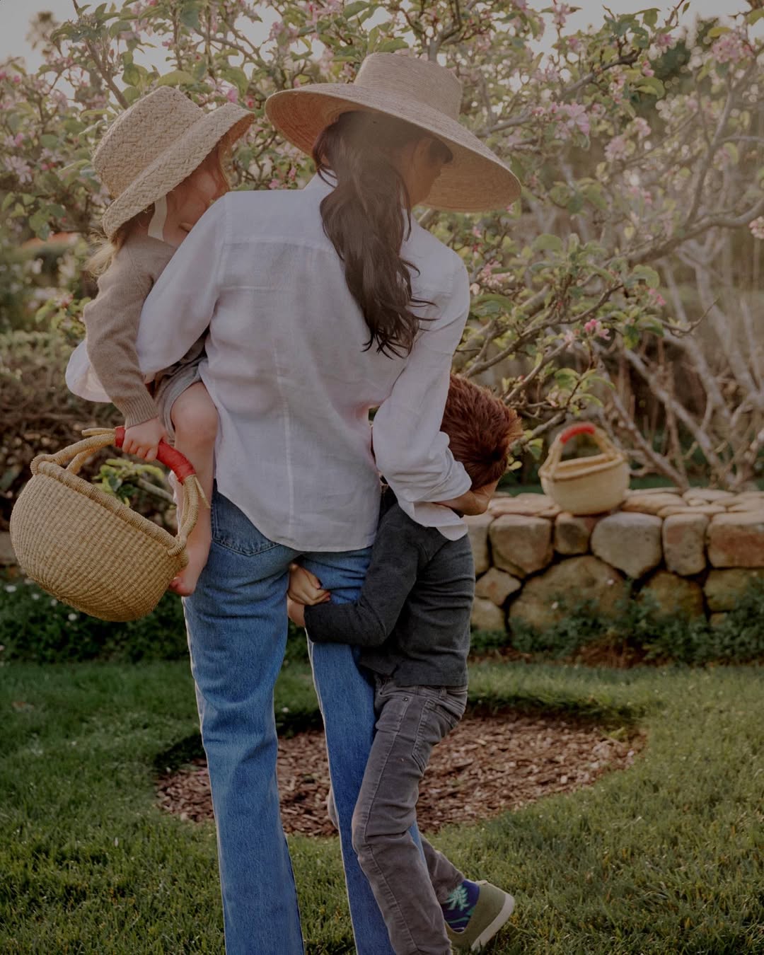 Meghan Markle walking in a garden carrying one child while another hugs her leg, both wearing straw hats.