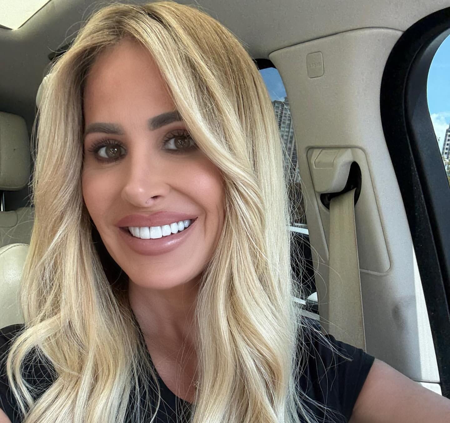 Kim Zolciak smiling in a car with long blonde hair, wearing a black top