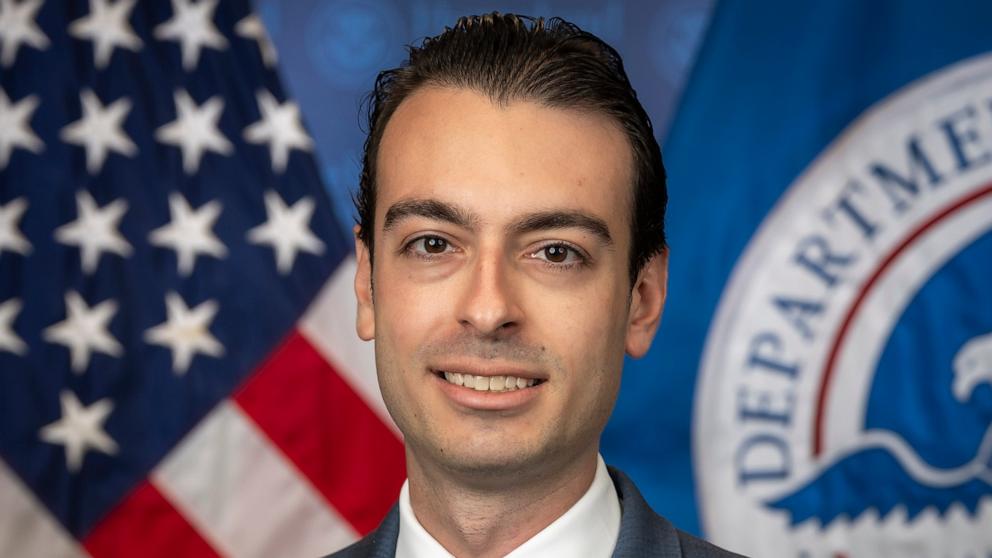 A man in a dark suit and white shirt poses in front of the American flag and a Department of Homeland Security emblem backdrop.