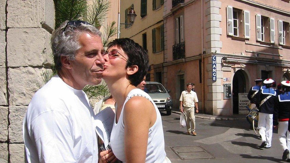 Jeffrey Epstein and Ghislaine Maxwell share a kiss on a street in a European town, with several people and uniformed guards walking nearby.