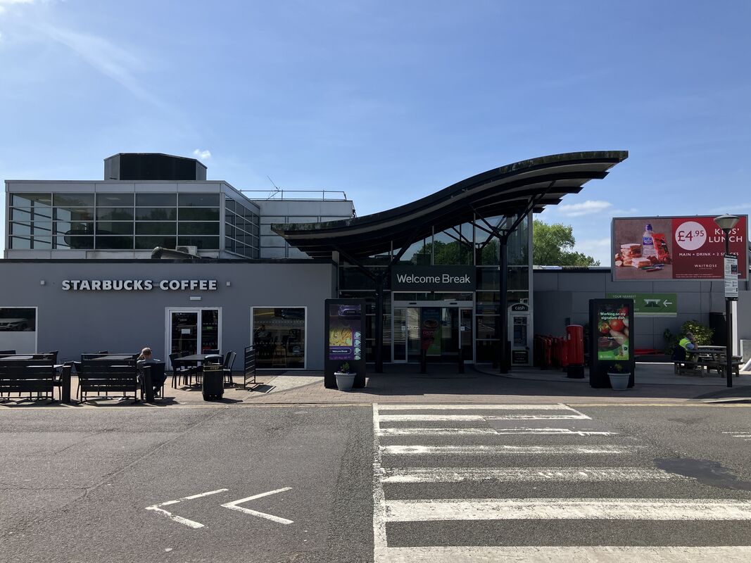 Exterior of Keele Services on the M6, showing the Welcome Break entrance and Starbucks seating area under clear blue skies.