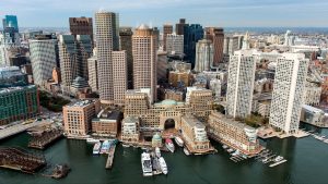 Aerial view of downtown Boston, Massachusetts, showing the Financial District skyline and Boston Harbor waterfront with boats docked along the piers.