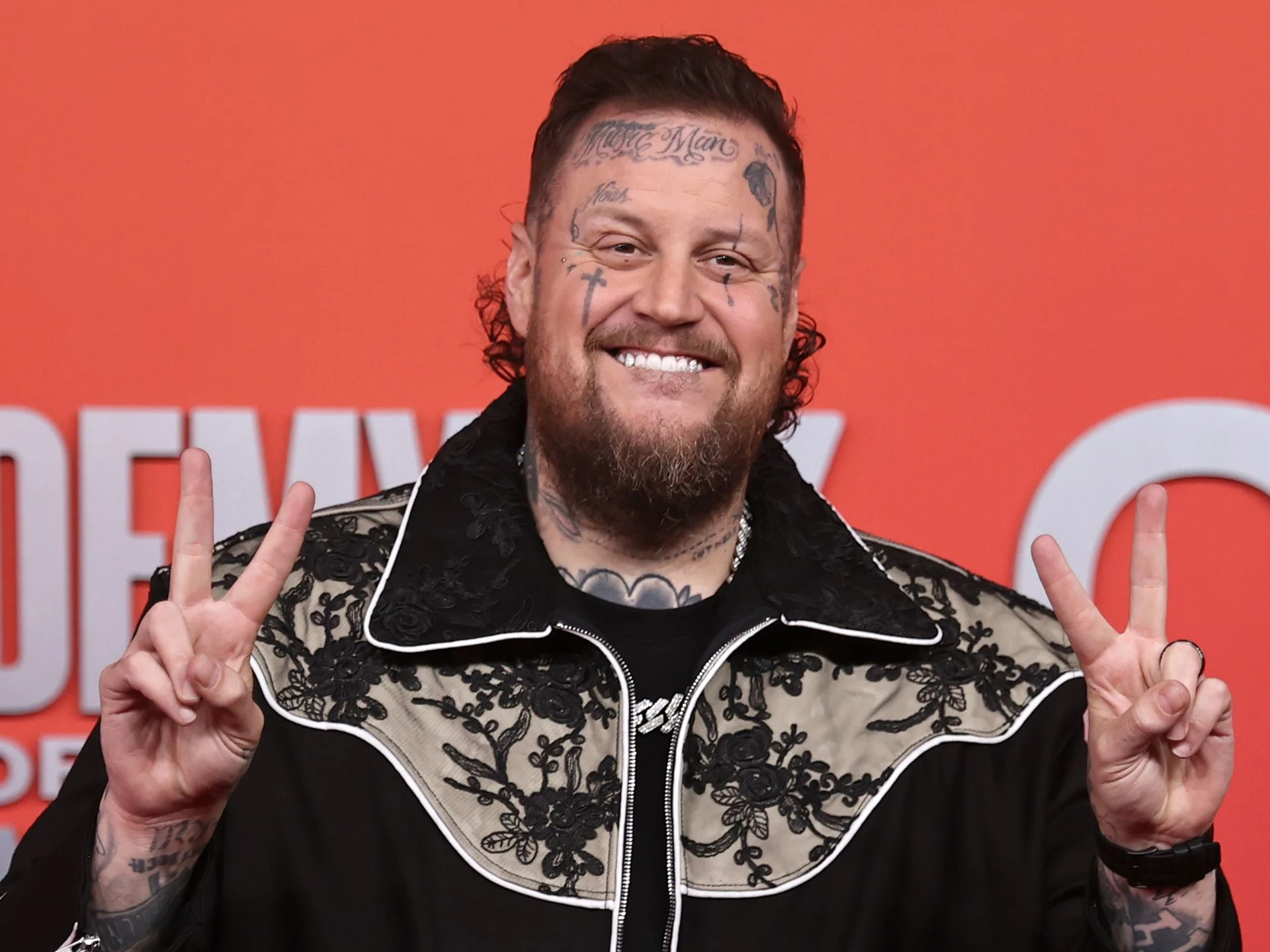 Jelly Roll smiles on the red carpet, flashing peace signs with both hands while wearing a black and white embroidered jacket.