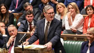A senior government official speaks at the despatch box during a tense parliamentary session, surrounded by MPs reacting to the debate.
