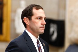 Rep. Jason Crow walking inside the U.S. Capitol, wearing a navy suit and striped tie, looking ahead with a serious expression.