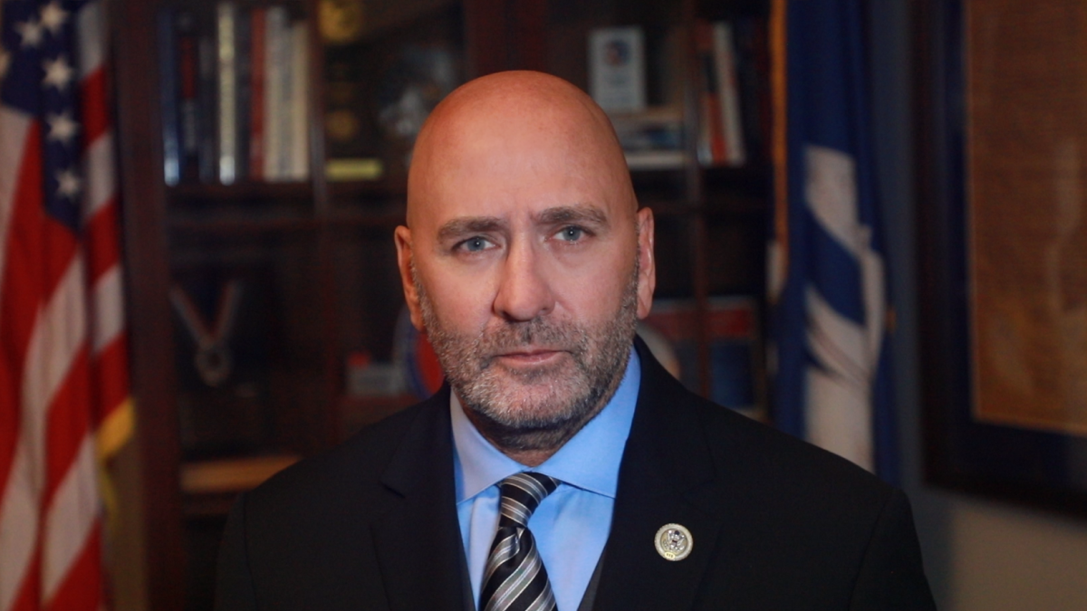 Rep. Clay Higgins stands in his office wearing a suit and tie, with U.S. flags and bookshelves visible in the background.