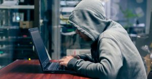 A person wearing a grey hoodie uses a laptop in a dimly lit room, symbolizing cybercrime or online harassment activity.