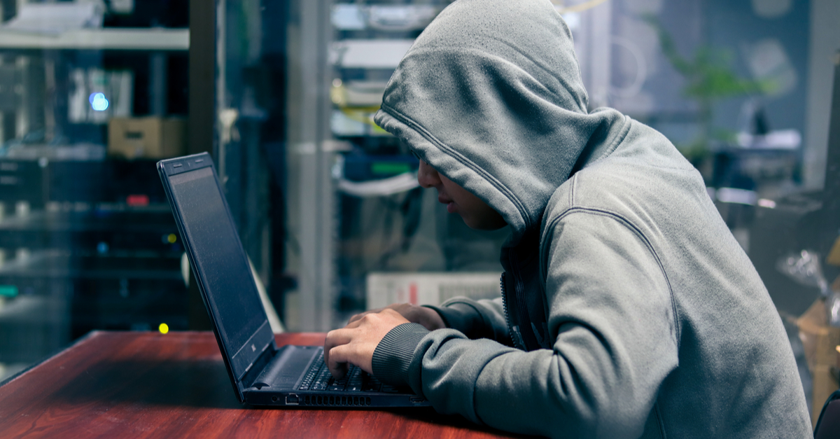 A person wearing a grey hoodie uses a laptop in a dimly lit room, symbolizing cybercrime or online harassment activity.
