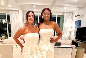 Kyle Richards and Garcelle Beauvais pose together wearing matching strapless white gowns with feather detailing inside a modern living room.