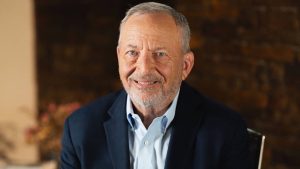 Larry Summers seated indoors, wearing a navy blazer and light blue shirt, looking toward the camera with a slight smile.