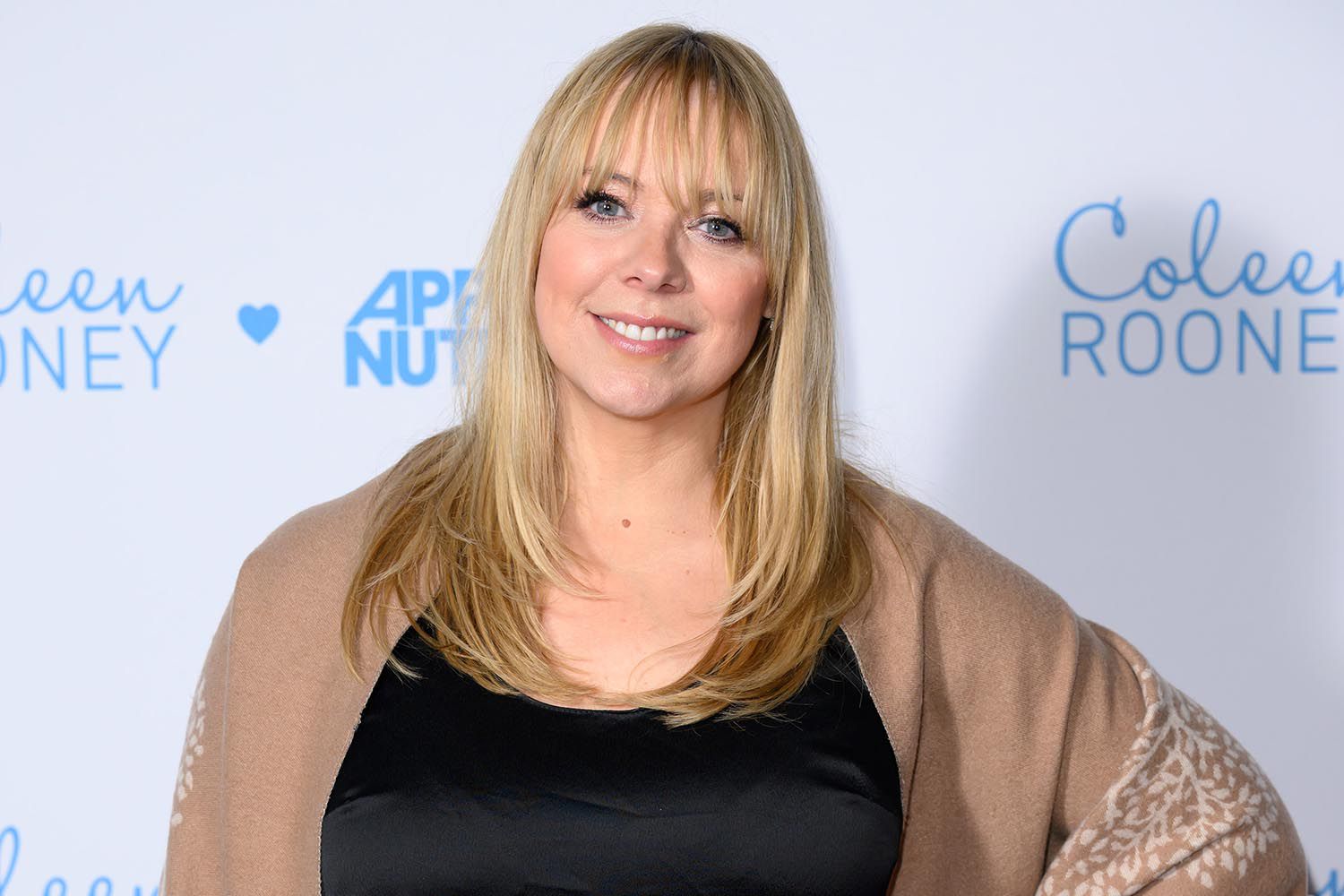 Liz McClarnon smiling on the red carpet wearing a black dress and beige shawl at a public event.