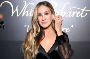 Sarah Jessica Parker attends a fashion event wearing a black satin wrap dress and diamond jewelry, posing with her hand to her ear.