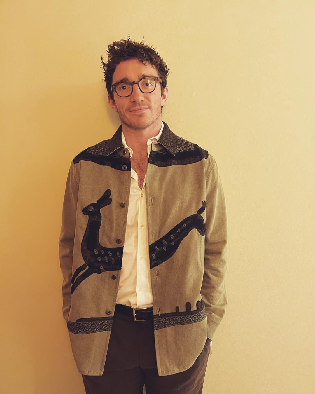 Caspar Jopling wearing a unique tan jacket with black embroidered deer and patterns, an unbuttoned white shirt, and glasses, standing against a yellow wall.