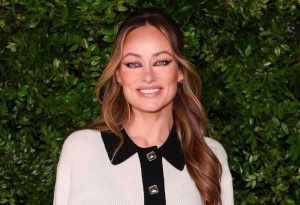 Olivia Wilde smiling in a cream and black collared sweater against a backdrop of lush green foliage.