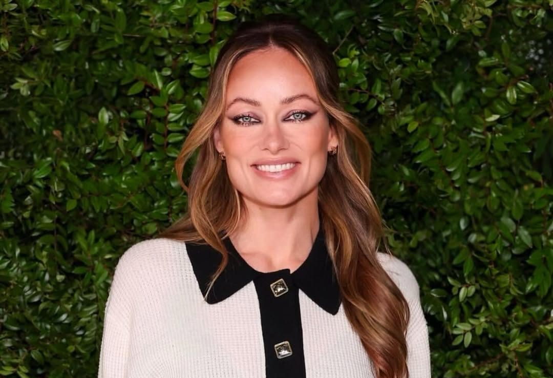 Olivia Wilde smiling in a cream and black collared sweater against a backdrop of lush green foliage.
