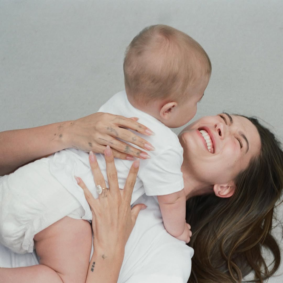 A smiling Hailey Bieber lying down and holding her infant son, Jack Blues Bieber, who is facing away from the camera.