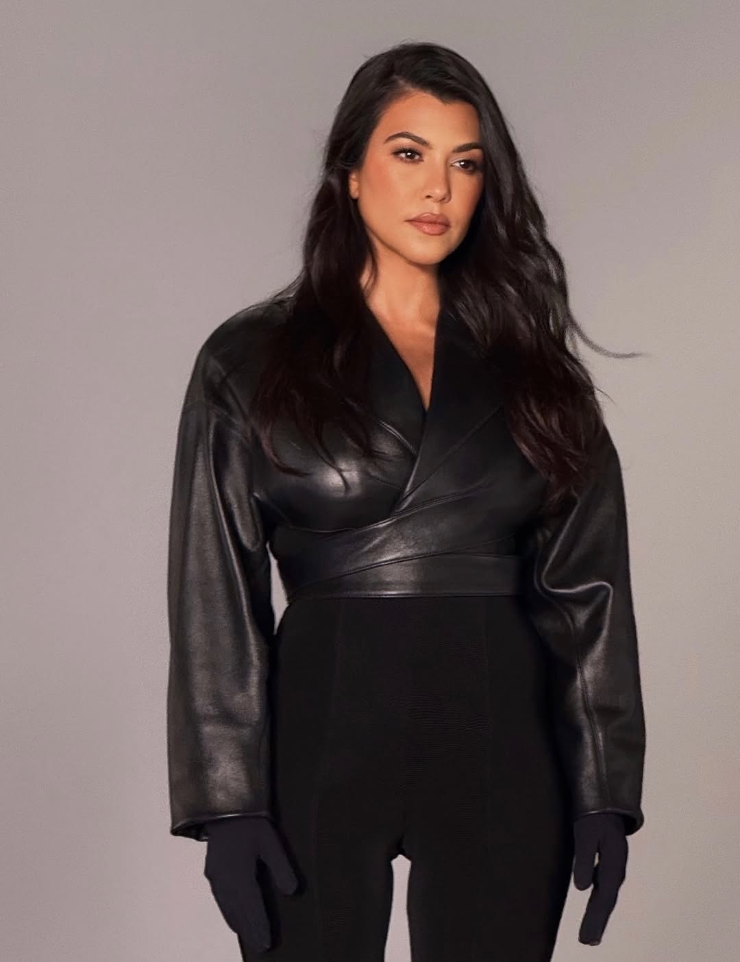 Studio portrait of Kourtney Kardashian wearing a black leather wrap jacket, black trousers, and long black gloves.