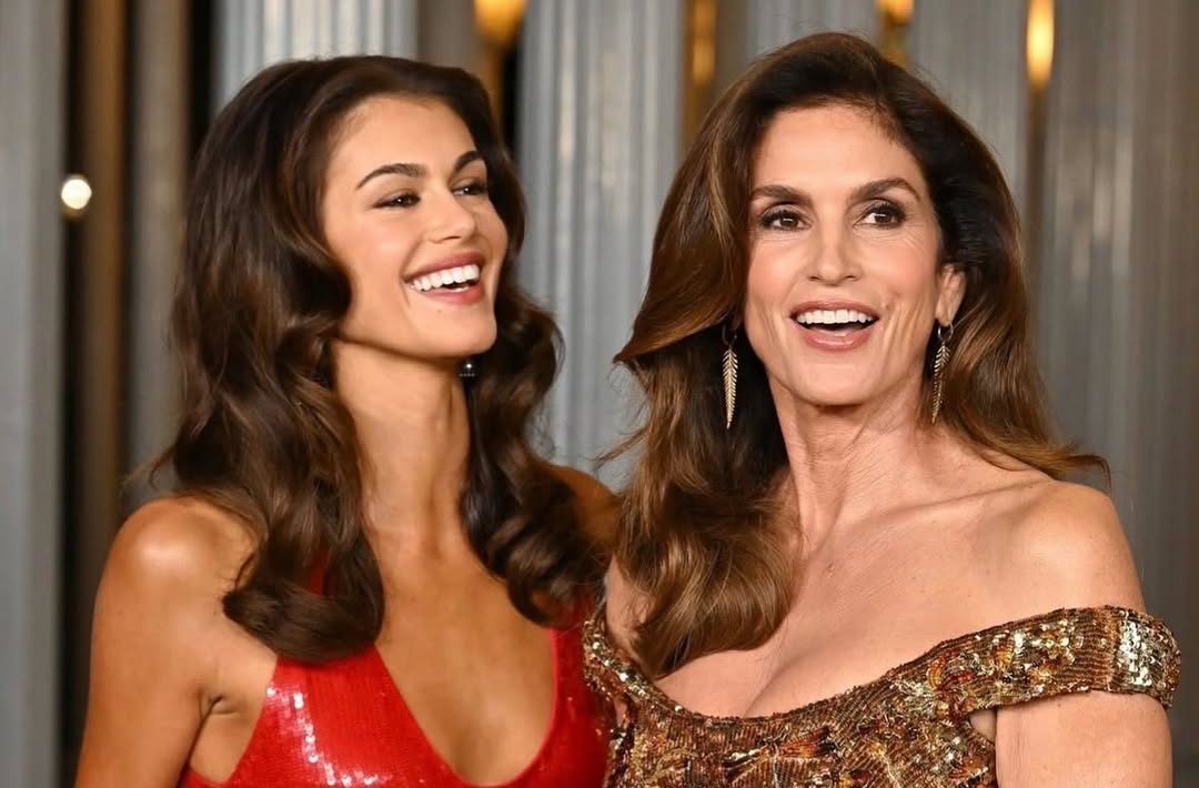 Kaia Gerber in a red sequined dress and Cindy Crawford in a gold sequined off-the-shoulder dress smiling at an event.