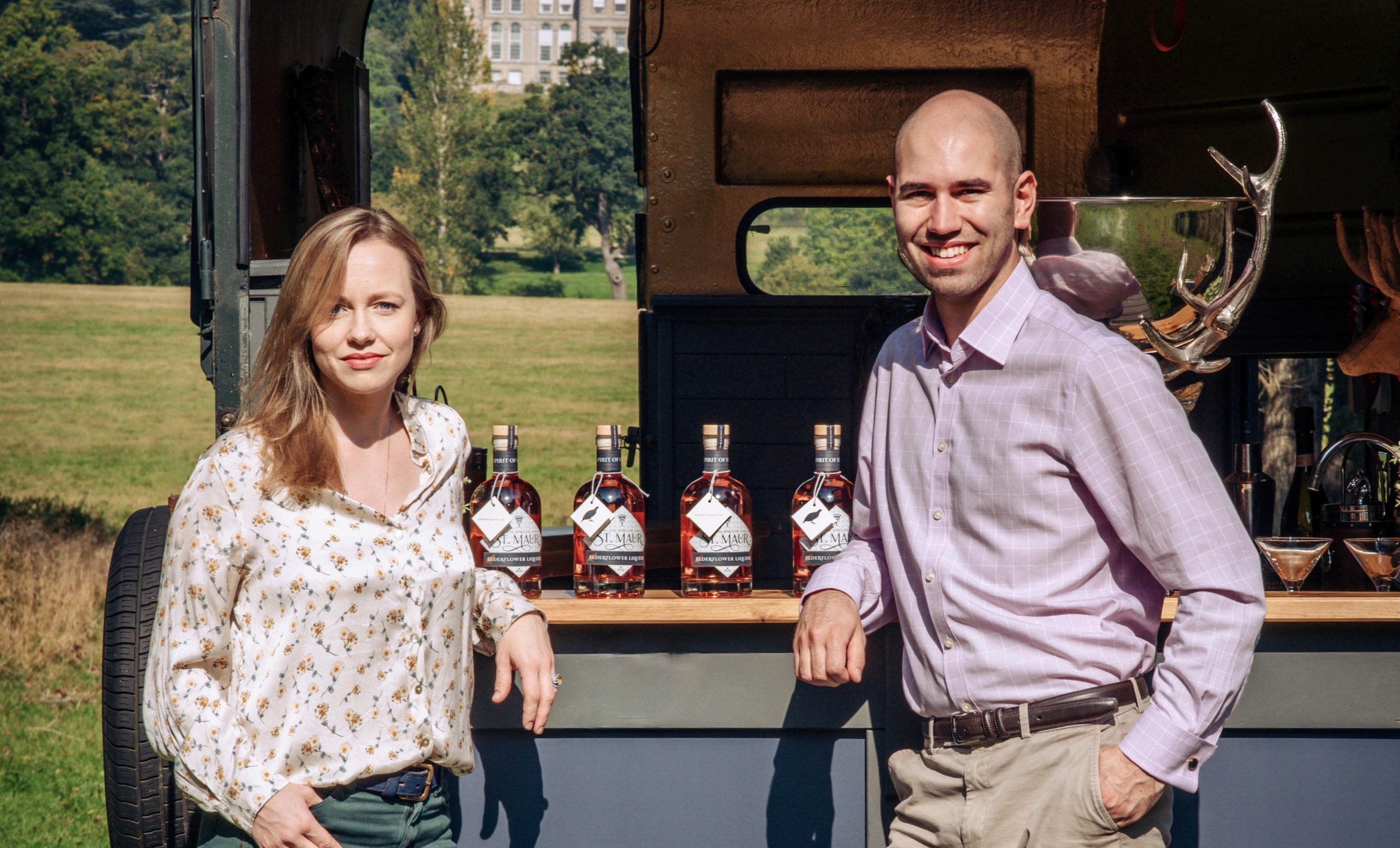 A man and woman stand beside a mobile bar featuring bottles of St Maur liqueur, posing outdoors in front of a scenic countryside estate.