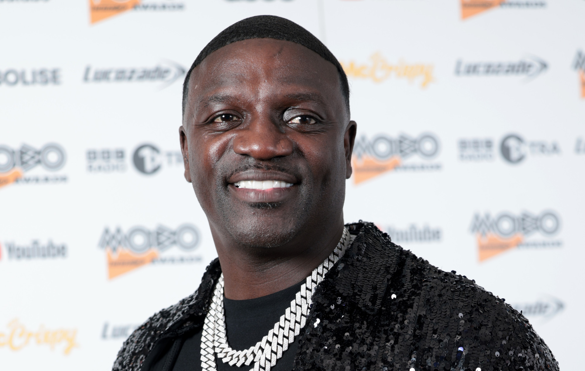 Akon attends the MOBO Awards in London wearing a black sequined jacket and diamond chain, smiling on the red carpet.