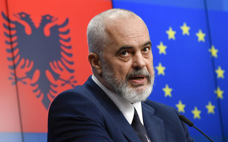Albanian Prime Minister Edi Rama speaking at a press conference in Brussels, with the Albanian and EU flags behind him.
