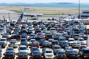 Crowded parking lot at Boston Logan Airport with rows of parked cars and airplanes taxiing on the runway in the background.