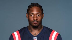 Headshot of NFL wide receiver Stefon Diggs, with cornrows, a mustache, and a goatee, wearing a dark blue New England Patriots jersey.