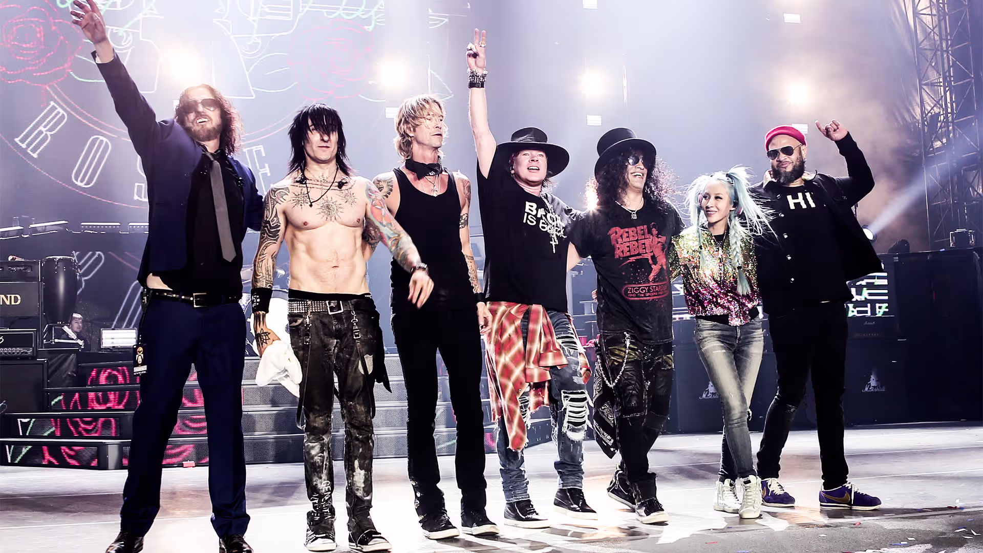 Guns N’ Roses band members take a bow on stage during a live concert performance, celebrating with the crowd under bright lights.