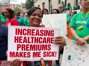 Protester holding a banner that reads “Healthcare premiums make me sick”