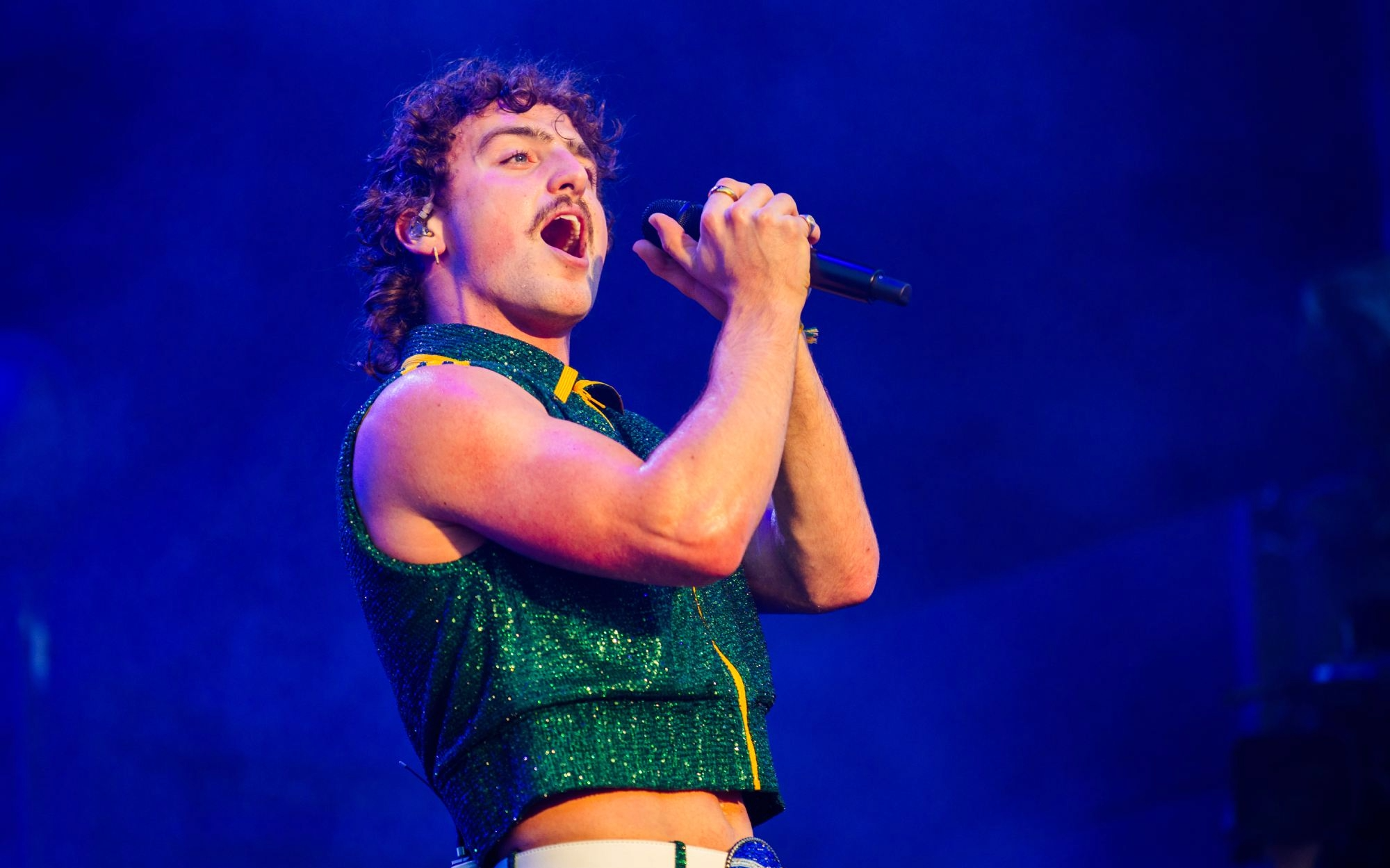 Singer performing energetically on stage under blue lighting, holding a microphone and wearing a sparkly green outfit during a live concert.