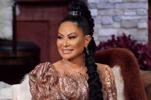 Jen Shah smiling during a televised reunion segment, wearing a sequined rose-gold dress and a long braided hairstyle, seated on a couch with a fur throw behind her.