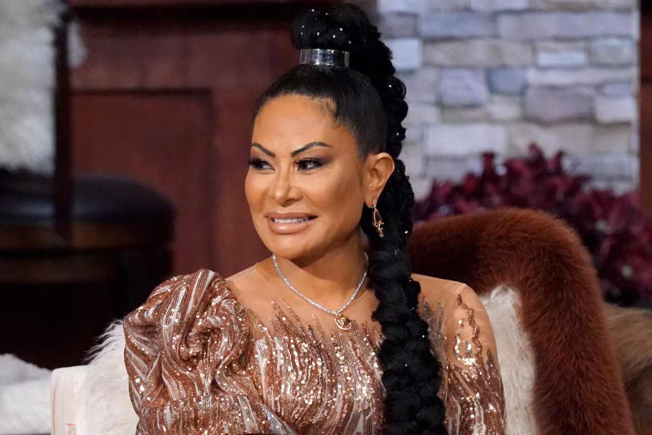 Jen Shah smiling during a televised reunion segment, wearing a sequined rose-gold dress and a long braided hairstyle, seated on a couch with a fur throw behind her.