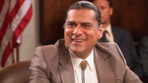 jonathan joss in a brown suit and bolo tie sits at a table in a formal meeting room with people seated behind him.